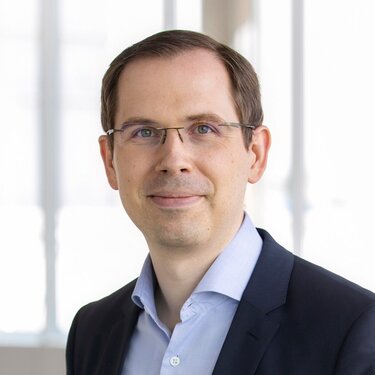 Ein Mann mit Brille und Anzug lächelt in die Kamera, vor einem hellen, unscharfen Hintergrund.