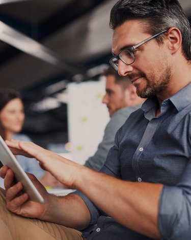 Ein Mann mit Brille sitzt in einem Büro und nutzt ein Tablet. Er trägt ein blaues Hemd und hat eine entspannte Körperhaltung. Im Hintergrund sind weitere Personen und eine Pinnwand sichtbar.