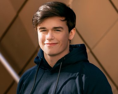 Junger Mann mit dunklem Kapuzenpullover steht lächelnd vor einer modernen, geometrischen Wand.