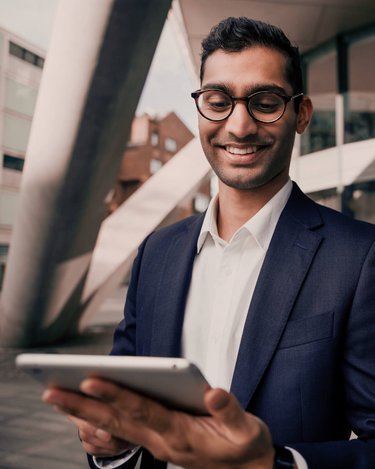 Ein Mann in einem Anzug hält ein Tablet und lächelt, während er vor einem modernen Gebäude steht. Die Architektur des Hintergrunds zeigt klare Linien und große Fenster.