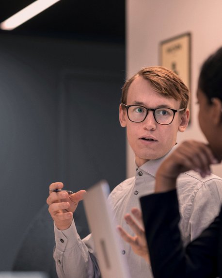 Ein Mann mit Brille spricht mit einer Frau in einem modernen Büro. Beide scheinen an einer Diskussion beteiligt zu sein.