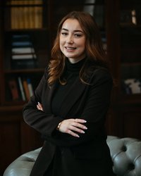 A woman with long brown hair stands with her arms crossed in an elegant black outfit in front of a bookshelf.