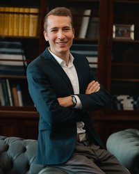 A young man in a dark blue suit is casually sitting on a sofa, with his arms crossed and a smiling expression on his face.