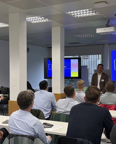 Ein Referent präsentiert vor einer Gruppe von Zuhörern in einem modernen Seminarraum. Eine Präsentation ist an der Wand sichtbar.