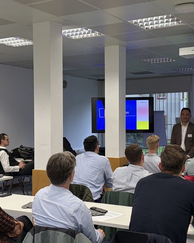 A speaker is presenting to a group of listeners in a modern seminar room. A presentation is visible on the wall.