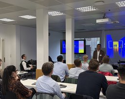 Ein Referent präsentiert vor einer Gruppe von Zuhörern in einem modernen Seminarraum. Eine Präsentation ist an der Wand sichtbar.