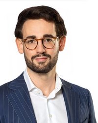 Mann mit Brille und Bart, trägt einen gestreiften Anzug und ein weißes Hemd, schaut direkt in die Kamera.