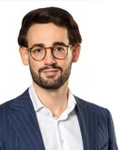 Mann mit Brille und Bart, trägt einen gestreiften Anzug und ein weißes Hemd, schaut direkt in die Kamera.