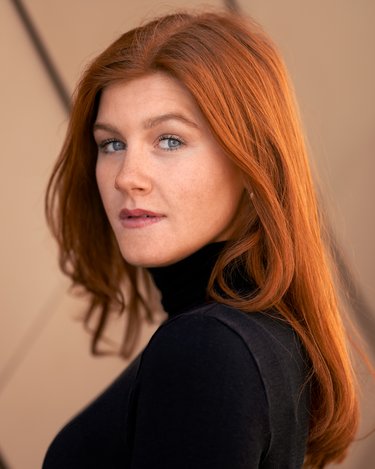 Eine Frau mit langen, roten Haaren und blauen Augen trägt einen schwarzen Rollkragenpullover und blickt über die Schulter.