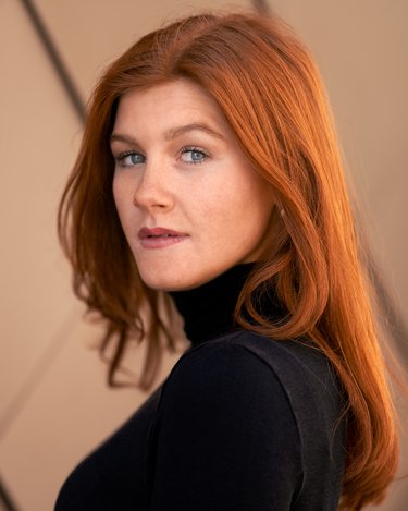 A woman with long red hair and blue eyes is wearing a black turtleneck and looking over her shoulder.