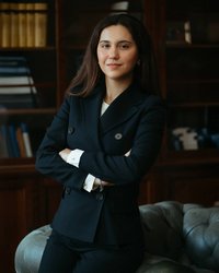 Young woman in a black blazer with her arms crossed, standing in front of a bookshelf and smiling slightly.