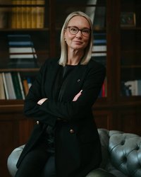 A woman with blonde hair and glasses stands with her arms crossed in an elegant black blazer in front of a bookshelf.
