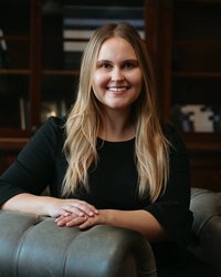 A smiling woman with long blonde hair is sitting in a green armchair, wearing a black top.