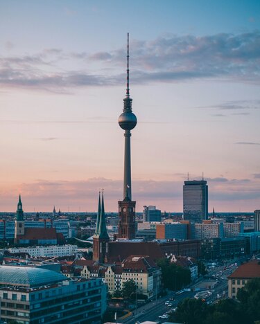 The Berlin TV Tower rises above the cityscape, surrounded by historic and modern buildings beneath a colorful sky.