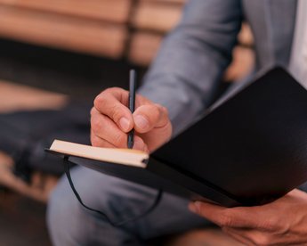 Eine Person in einem Anzug hält einen Stift und schreibt in ein schwarzes Notizbuch.