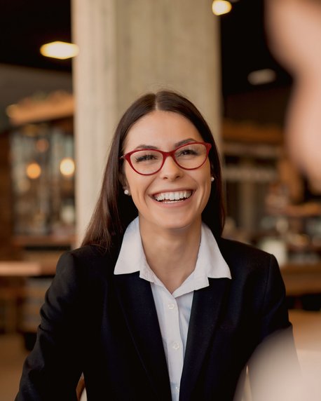 Eine Frau mit langen, dunklen Haaren und roten Brillenläsern lächelt in einem Café. Sie trägt einen schwarzen Blazer und sitzt an einem Tisch, während sie mit zwei anderen Personen spricht.
