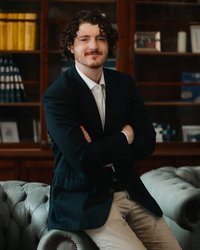 Young man with curly hair sitting on a sofa in an elegant suit, arms crossed. Bookshelf in the background.