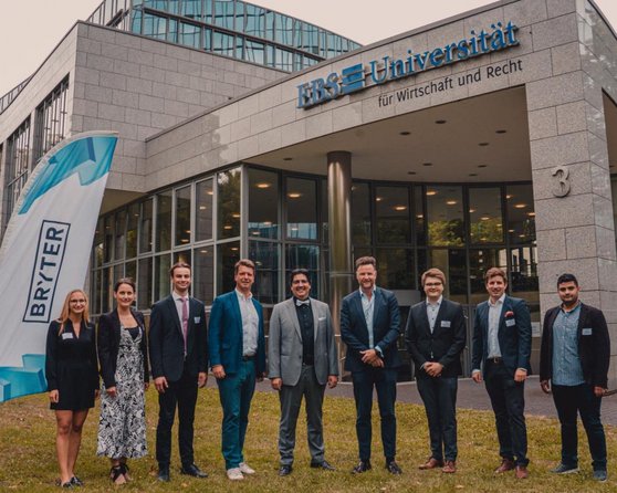 Group of nine people in front of the University of Applied Sciences for Business and Law, holding two flags displaying the 'BRYTER' logo. The individuals are dressed in formal attire and are standing in a line.