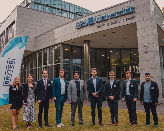 Gruppierung von neun Personen vor dem Gebäude der Hochschule für Wirtschaft und Recht, mit zwei Fahnen, die das Logo von "BRYTER" zeigen. Die Personen tragen formelle Kleidung und stehen in einer Reihe.