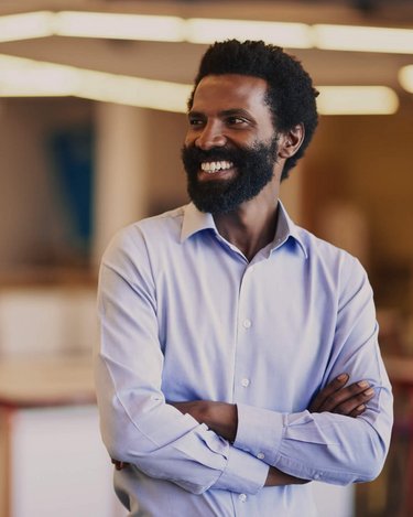 Ein Mann mit Bart und lockigem Haar trägt ein hellblaues Hemd und steht in einem modernen, hellen Raum mit Tischen und Stühlen. Er hat die Arme verschränkt und lächelt.