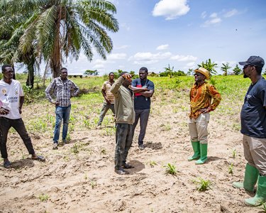 A group of six people stands in a field while one person speaks. Some are wearing rubber boots and discussing agricultural topics.