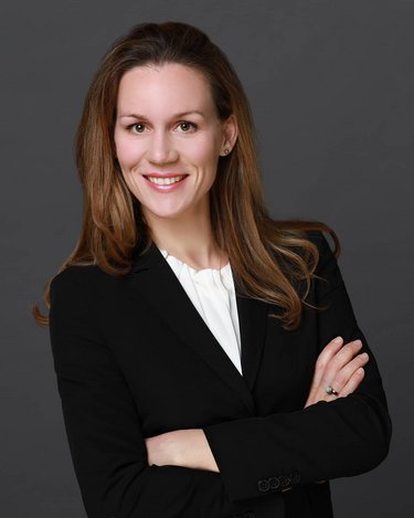 Eine Frau mit langen, braunen Haaren trägt einen schwarzen Blazer und lächelt, während sie mit verschränkten Armen vor einem grauen Hintergrund steht.