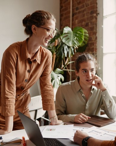 Three people in an office are discussing documents and notes while a laptop sits on the table. Plants are visible in the background.