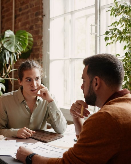 Drei Personen in einem Büro diskutieren über Dokumente und Notizen, während ein Laptop auf dem Tisch steht. Pflanzen sind im Hintergrund sichtbar.