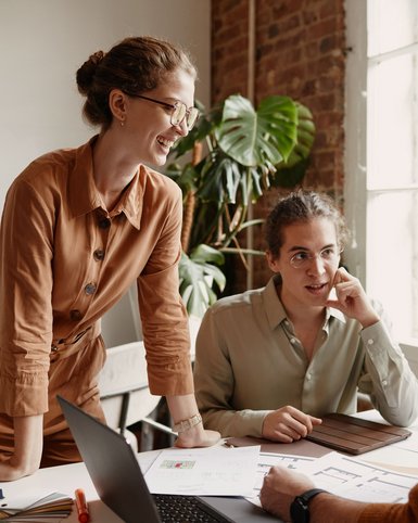 Drei Personen in einem Büro diskutieren über Dokumente und Notizen, während ein Laptop auf dem Tisch steht. Pflanzen sind im Hintergrund sichtbar.