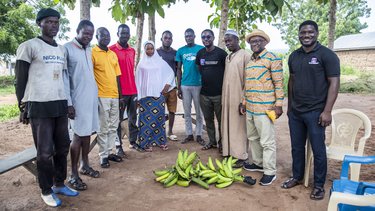 Eine Gruppe von neun Personen steht um einen Haufen Bananen, umgeben von Bäumen und einem ländlichen Hintergrund.
