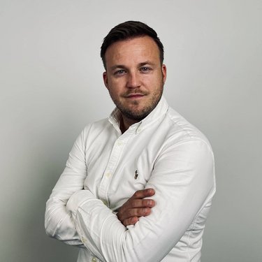 Mann mit kurzen Haaren und Bart, trägt ein weißes Hemd, steht mit verschränkten Armen vor einer neutralen Wand.