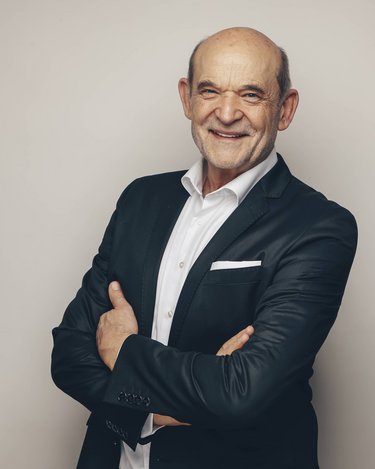 Ein älterer Mann mit Glatze und breitem Lächeln, gekleidet in einem schwarzen Anzug mit weißem Hemd, steht mit verschränkten Armen vor einem neutralen Hintergrund.