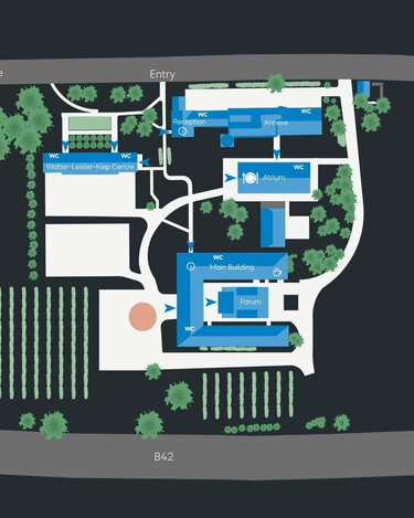 Site plan of an area featuring various buildings, entrances, green spaces, and a parking lot. Labels indicate important facilities such as restrooms and the main building.
