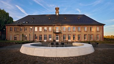 A historic building with a clock tower, large windows, and a modern, round seating area in the foreground. The sky is clear with a few clouds, and the surroundings are calm and inviting.
