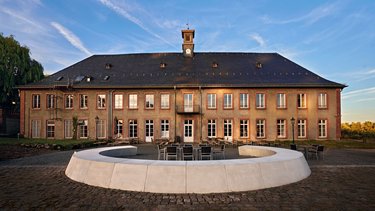 Ein historisches Gebäude mit einem Uhrenturm, großen Fenstern und einem modernen, runden Sitzbereich im Vordergrund. Der Himmel ist klar mit einigen Wolken, und die Umgebung ist ruhig und einladend.