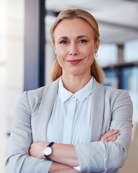 Eine Frau mit blonden Haaren und einem grauen Blazer steht mit verschränkten Armen in einem modernen Büro.