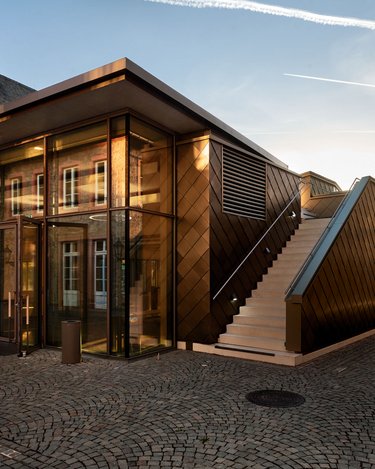 Ein modernes Gebäude mit einer Glasfassade und einer schrägen Treppe, umgeben von einem gepflasterten Platz. Die Architektur kombiniert zeitgenössische Elemente mit traditionellen Materialien.