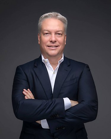 Man in a dark blue suit with crossed arms, smiling against a neutral background.