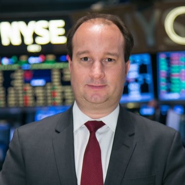 Mann in Anzug mit Krawatte steht vor einem Bildschirm mit Börsendaten. Hintergrund zeigt die NYSE.