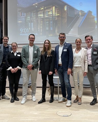A group of eight people stands on a stage, smiling, dressed in formal attire, with a screen in the background.