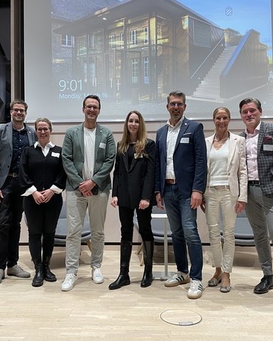 Eine Gruppe von acht Personen steht auf einer Bühne, lächelnd, in formeller Kleidung, mit einem Bildschirm im Hintergrund.