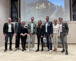 Eine Gruppe von acht Personen steht auf einer Bühne, lächelnd, in formeller Kleidung, mit einem Bildschirm im Hintergrund.