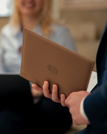 Ein Mann hält eine braune Mappe in der Hand, während zwei Personen im Hintergrund an einem Laptop sitzen und miteinander sprechen.