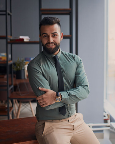 A man with a beard is wearing a green shirt and a black tie as he casually leans against a table in a modern room. In the background, shelves and windows are visible.