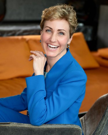 Frau mit kurzen, blonden Haaren in einem blauen Anzug sitzt lächelnd auf einem Stuhl, mit orangefarbenem Hintergrund.