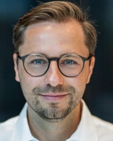 Ein Mann mit Brille und kurzem, gestyltem Haar, der direkt in die Kamera schaut. Er trägt ein weißes Hemd und hat einen gepflegten Bart.