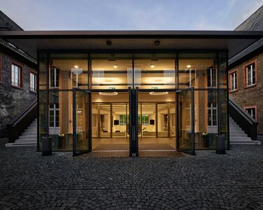 Modern glass doors lead into a bright foyer, surrounded by historic buildings with brick walls and windows.
