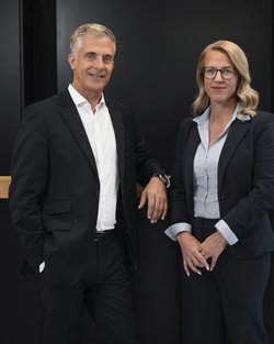 A man and a woman are standing next to each other at a reception desk. Both are dressed in formal attire and smiling.