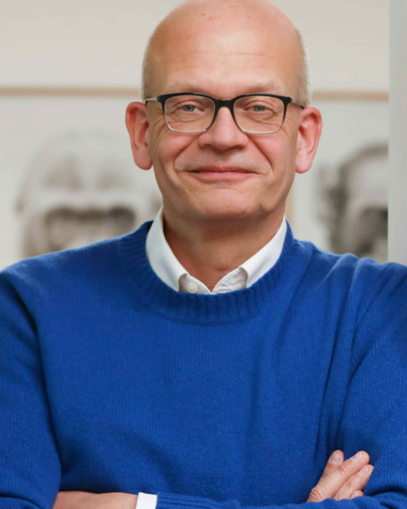 Ein Mann mit Brille und einem blauen Pullover steht mit verschränkten Armen und lächelt. Im Hintergrund sind schemenhafte Zeichnungen zu sehen.