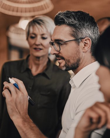 Ein Mann mit Brille schreibt an einer Tafel, während zwei Frauen ihm aufmerksam zuschauen. Die Szene spielt sich in einem modernen Büro oder Besprechungsraum ab.